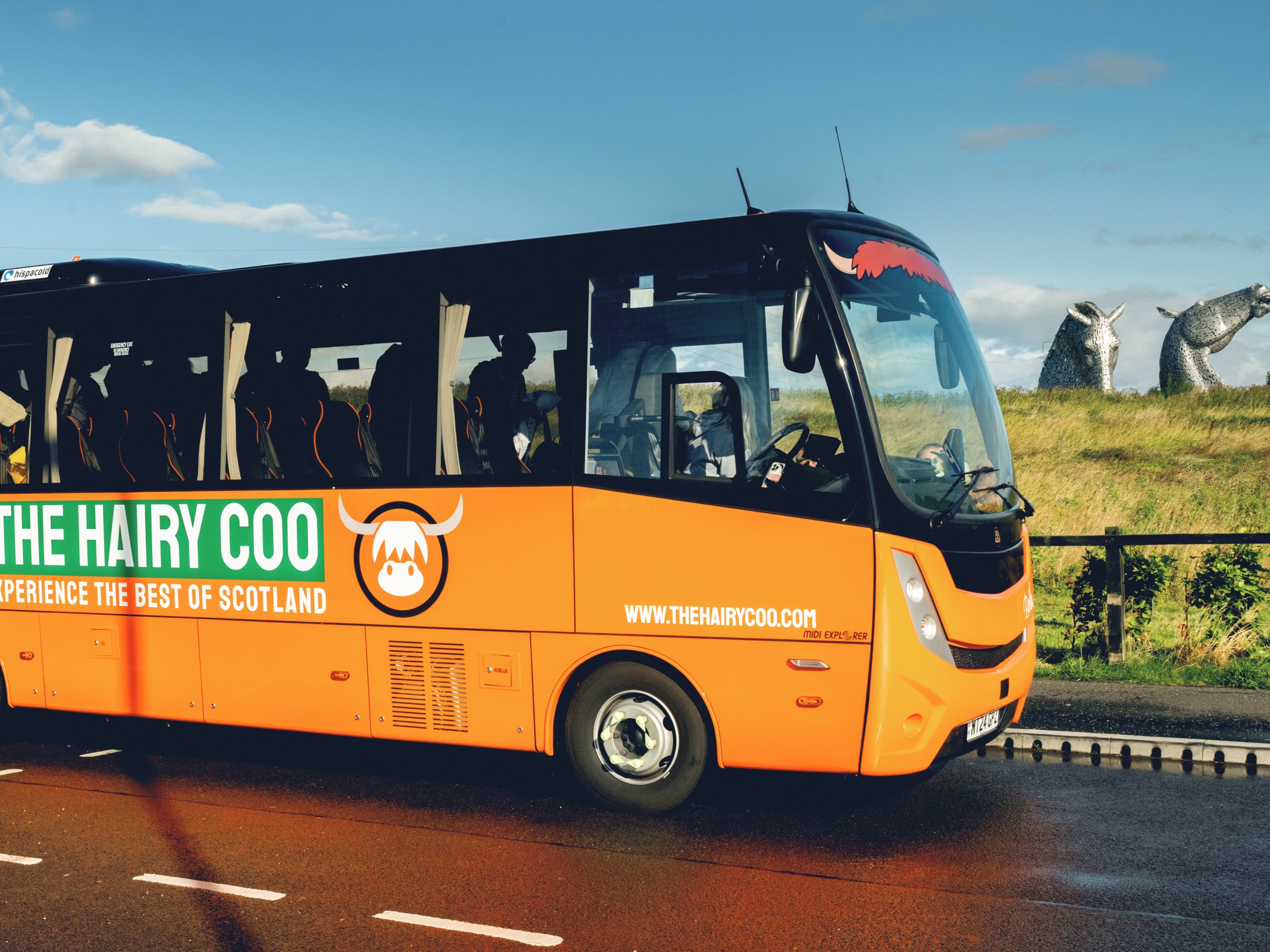 A Hairy Coo orange coach with the Kelpies in the background