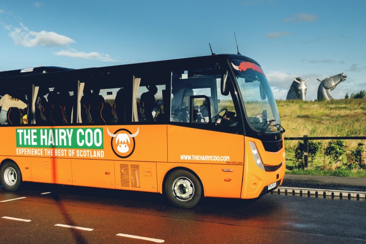 A Hairy Coo orange coach with the Kelpies in the background