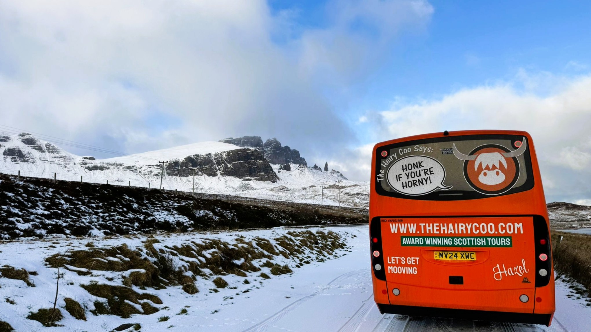 Orange tour bus on snowy road on Isle of Skye. The Hairy Coo bus with mountain backdrop under cloudy sky.