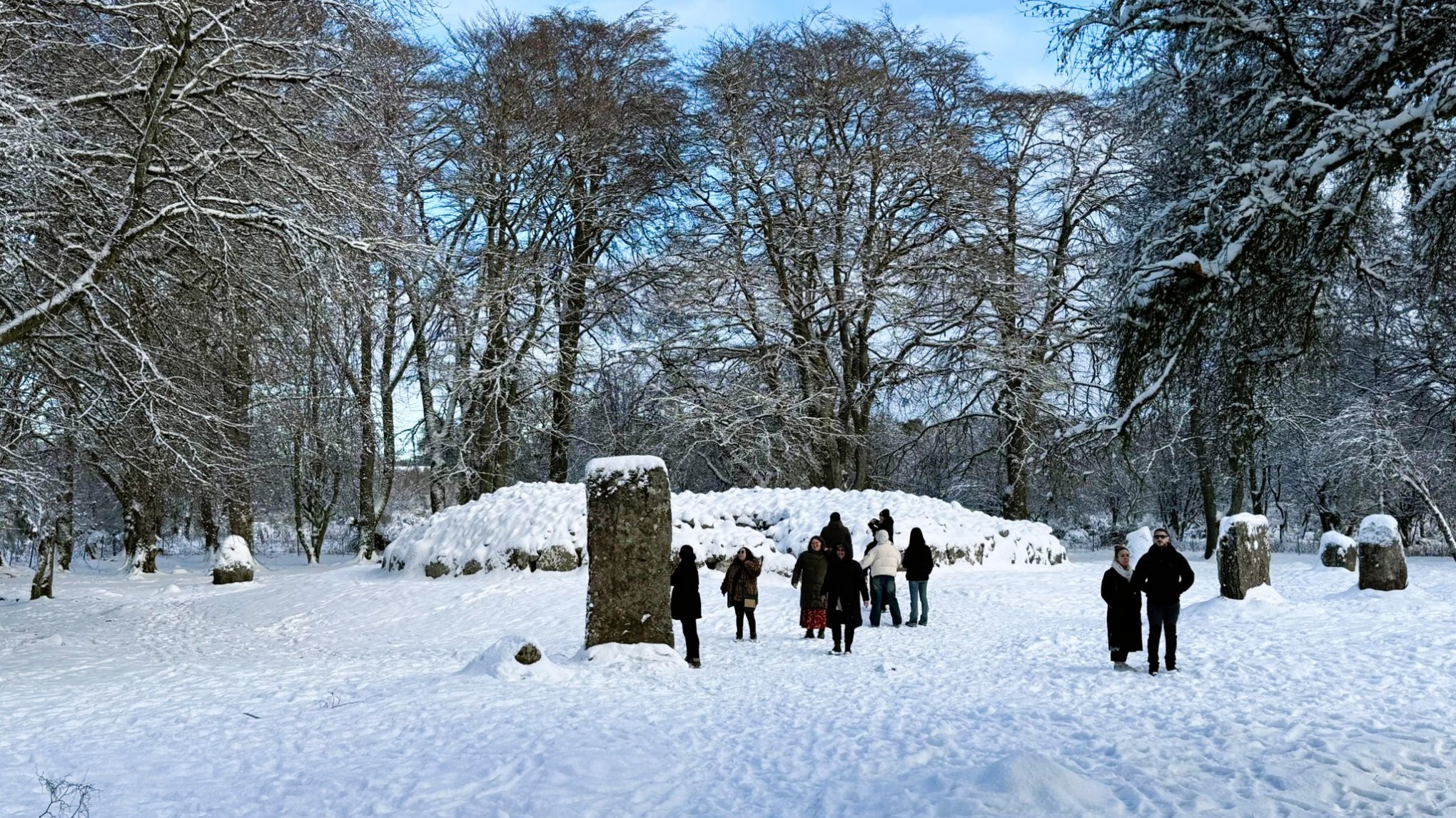 People exploring Clava Cairns in the snow with standing stones, trees covered in snow.